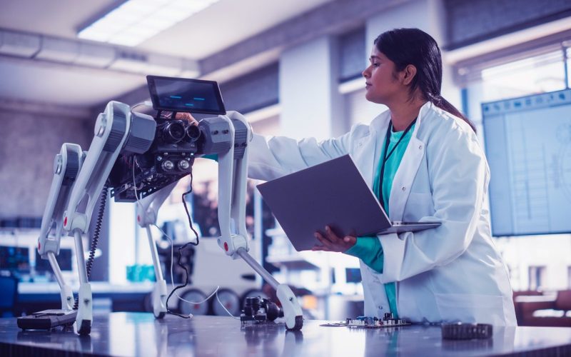 Young Indian Female Engineer Testing Industrial Programmable Robot Animal in a Factory Development Workshop. Professional Researcher in a Lab Coat Developing AI Canine Prototype, Using Laptop Computer